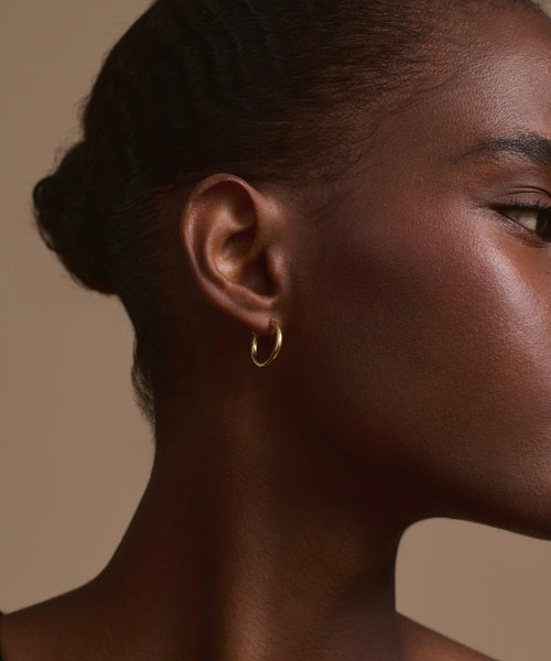 Close-up of the right side of a woman’s face and ear, highlighting her smooth skin, hair pulled back, and a Small Eloise Hoop earring by Jenni Kayne in 14k gold. The background is solid beige.