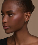 A woman with dark skin and short, pulled-back hair looks left, wearing the Small Eloise Hoops and a black top against a neutral beige background.