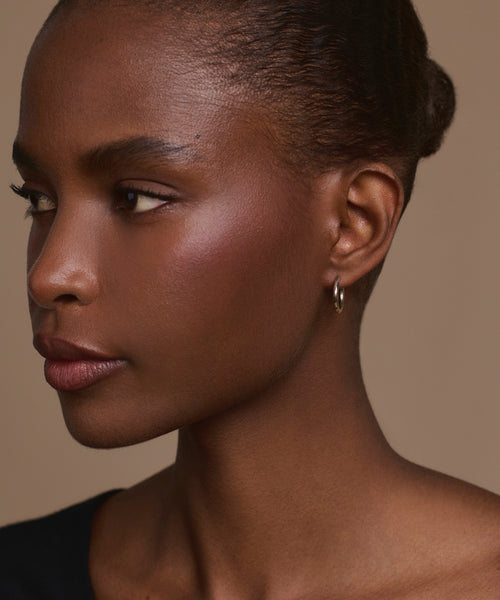 A woman with dark skin and short, pulled-back hair looks left, wearing the Small Eloise Hoops and a black top against a neutral beige background.