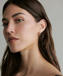 A woman with long brown hair wears Jenni Kayne’s Small Eloise Hoops in 14k gold, looking slightly to the side with a calm, neutral expression against a plain background.