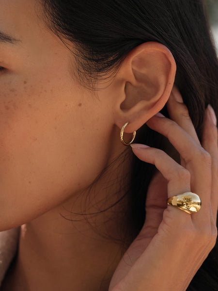 Close-up of a person’s ear wearing Small Eloise Hoops by Jenni Kayne. Their manicured hand touches the ear, highlighting a chunky gold ring—an elegant mix of fine jewelry against freckled skin.