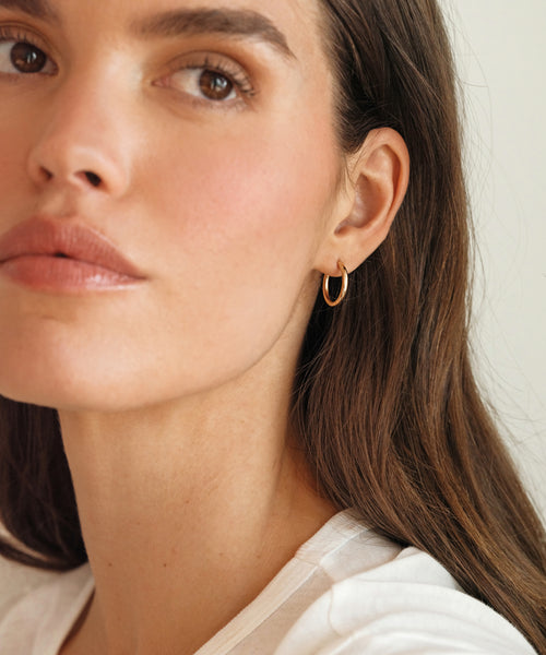 A woman with long brown hair, wearing a white top and the Small Eloise Hoops by Jenni Kayne, gazes calmly at the camera against a neutral background.