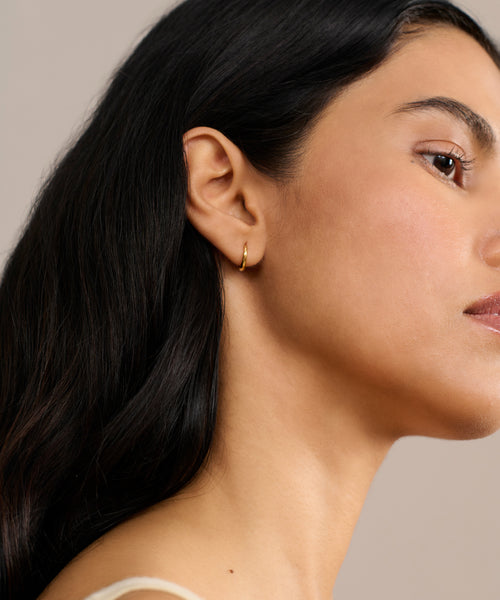 A woman with long dark hair is shown in profile against a neutral background, wearing a cream top and Small Hellenic Hoops—handcrafted 18k gold vermeil earrings made in Los Angeles. Only the right side of her face and shoulder are visible.