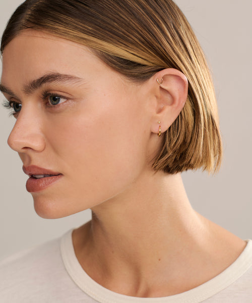 A person with short, straight light brown hair wears Small Hellenic Hoops—18k gold vermeil earrings—and a white shirt, gazing slightly left against a neutral background.