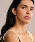 A woman with long dark hair and radiant skin looks confidently at the camera, wearing a sleeveless beige top, natural makeup, and Small Hellenic Hoops—18k gold vermeil earrings handcrafted in Los Angeles—against a neutral background.