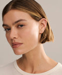 A woman with light skin and short, straight brown hair looks slightly off camera; she wears Small Hellenic Hoops—handcrafted 18k gold vermeil earrings made in Los Angeles—and a white crew neck top.