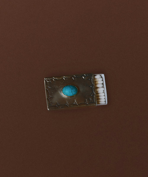 The Small Stamped Matchbox with Turquoise features a metallic embossed cover and a blue turquoise stone, set against a brown background.