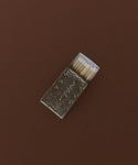 A Small Silver Stamped Matchbox with a metallic, textured cover rests on a brown surface, partially open to reveal several wooden matches with white tips inside.