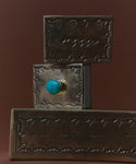 Three Small Silver Stamped Matchboxes with intricate designs are stacked. The middle one, made by a Navajo silversmith, features a central round turquoise stone. The background is a plain, dark brown color.