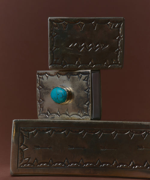 Three Small Silver Stamped Matchboxes with intricate designs are stacked. The middle one, made by a Navajo silversmith, features a central round turquoise stone. The background is a plain, dark brown color.