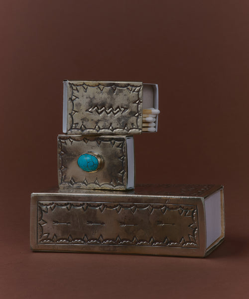 Three decorative matchboxes are stacked on a brown surface; one is the Small Silver Stamped Matchbox, featuring a turquoise knob, while another matchbox is partially open to reveal matches inside.