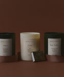 Three scented candles labeled Aspen, Cedar, and Sun Valley by Jenni Kayne are displayed against a brown background, with a Small Silver Stamped Matchbox and a ribbon placed in front.