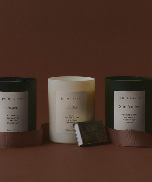 Three scented candles labeled Aspen, Cedar, and Sun Valley by Jenni Kayne are displayed against a brown background, with a Small Silver Stamped Matchbox and a ribbon placed in front.