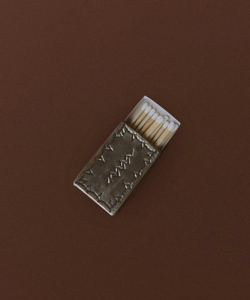 A Small Silver Stamped Matchbox with a metallic, textured cover rests on a brown surface, partially open to reveal several wooden matches with white tips inside.
