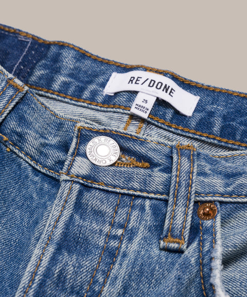 Close-up view of the waistband and button of The Standard blue denim straight leg jeans. The brand label reads RE/DONE, size tag 25. Inspired by Men’s 501 Levi’s, they feature gold stitching and an “Original” metal button.