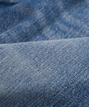 Close-up of blue denim fabric with a textured weave and subtle color variations. Soft folds create a wavy look, reflecting the classic style of The Standard jeans with a higher mid-rise and straight leg design.