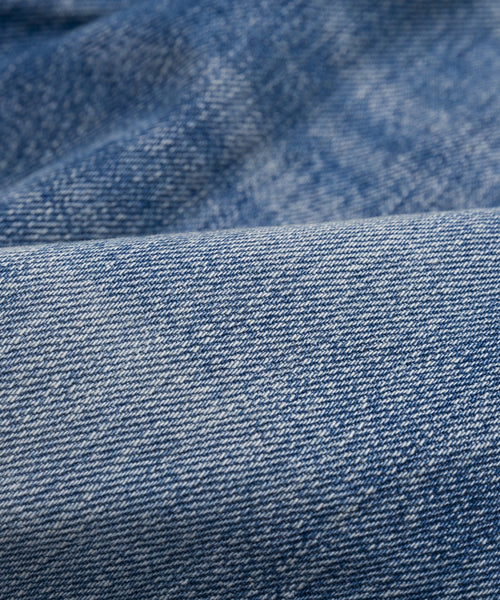 Close-up of blue denim fabric with a textured weave and subtle color variations. Soft folds create a wavy look, reflecting the classic style of The Standard jeans with a higher mid-rise and straight leg design.