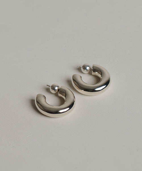 The Large Donut Hoops are sterling silver earrings featuring thick, rounded bands and small spherical studs, highlighting sculptural design on a simple, light backdrop.