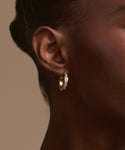 Close-up of a woman's ear wearing Stevie Grey Diamond Hoops, featuring sparkling gemstones. The plain beige background accentuates the stunning heirloom jewelry and her smooth, dark skin tone.