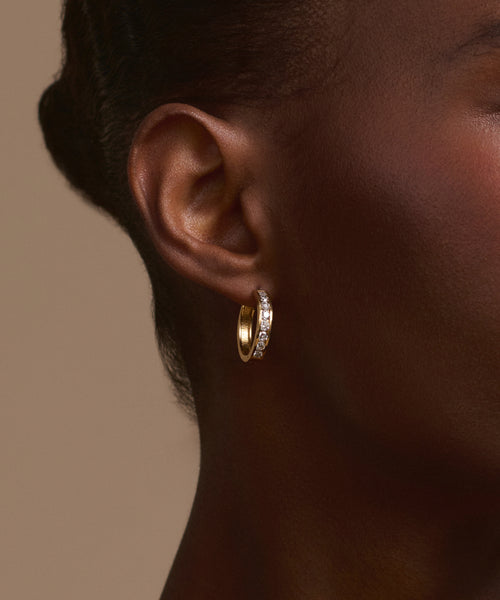 Close-up of a woman's ear wearing Stevie Grey Diamond Hoops, featuring sparkling gemstones. The plain beige background accentuates the stunning heirloom jewelry and her smooth, dark skin tone.