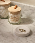Two Stowe Eco Glass Canisters with wooden lids, one holding cotton balls and the other cotton swabs, rest on a marble countertop next to a white ring dish—an elegant and sustainable storage solution.