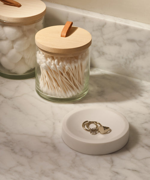 Two Stowe Eco Glass Canisters with wooden lids, one holding cotton balls and the other cotton swabs, rest on a marble countertop next to a white ring dish—an elegant and sustainable storage solution.