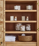 A wooden cabinet with shelves displaying candles, skincare items, jars of cotton balls and pads, the Stowe Eco Glass Canister for cotton swabs, folded white towels, and a woven basket for sustainable storage.