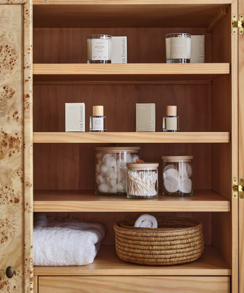 A wooden cabinet with shelves displaying candles, skincare items, jars of cotton balls and pads, the Stowe Eco Glass Canister for cotton swabs, folded white towels, and a woven basket for sustainable storage.