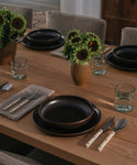 A dining table set with ceramic dinnerware and recycled glass cups with vases filled with florals. 