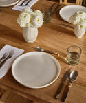 A wooden dining table is set with white plates, silver cutlery, Straight Rock Glass tumblers filled with water, white cloth napkins, and two white vases holding white ranunculus flowers.
