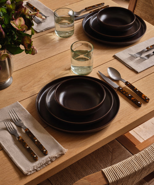 A dining table set with ceramic dinnerware and recycled glass cups with vases filled with florals. 