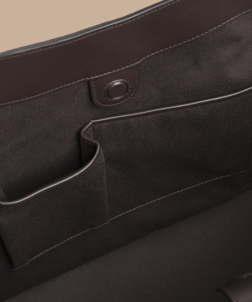 Close-up of the inside of a dark brown Suede Agnes Tote, featuring two fabric pockets and a snap button closure on a leather flap—ideal for use as a laptop bag.