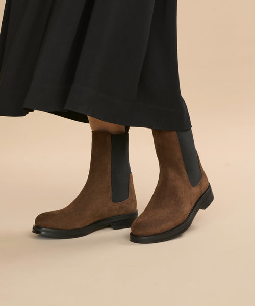 A person in a long black skirt wears the Suede Alden Boot—brown suede Chelsea boots with black elastic side panels and a weather-friendly rubber sole—standing on a light beige background.