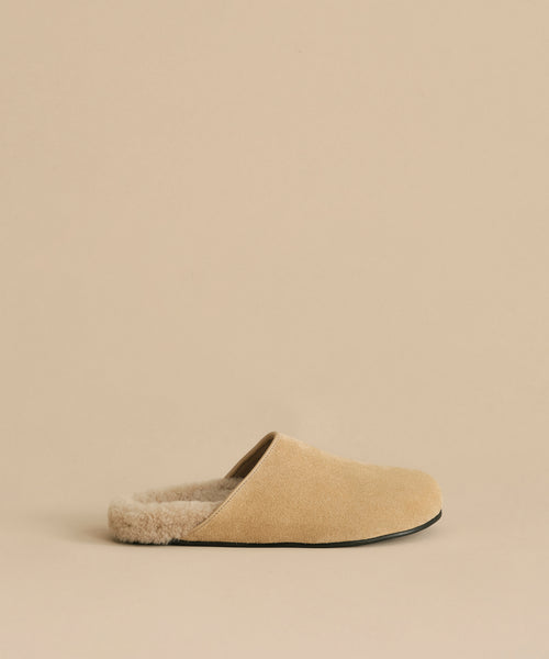 The Suede Ashby Clog features tan suede, a fluffy shearling-lined footbed, and a black sole, shown on a matching beige background.