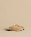 The Suede Ashby Clog features tan suede, a shearling-lined footbed, plush furry lining, and black soles, shown here on a matching beige background.