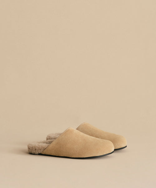 The Suede Ashby Clog features tan suede, a shearling-lined footbed, plush furry lining, and black soles, shown here on a matching beige background.