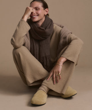 A person sits cross-legged on the floor, smiling with one hand on their face. They wear beige loungewear, a brown scarf, gold jewelry, and the Suede Ashby Clog—featuring a cozy shearling-lined footbed—against a beige background.
