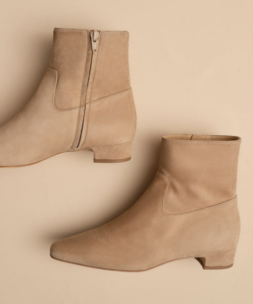 The Suede Carmine Boot features taupe suede, low heels, side zippers, and a minimalist design, shown against a matching beige background.