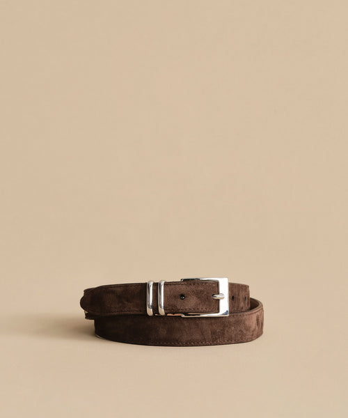 A Suede Danner Belt in brown, featuring a shiny silver buckle, is coiled neatly on a plain beige background, highlighting its timeless elegance.