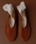 The Suede Elodie Flat, crafted in Italy with a feminine silhouette, is styled with white ribbed socks and displayed elegantly on a brown surface.
