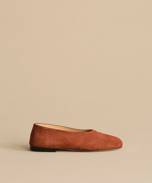 The Suede Elodie Flat, featuring a feminine silhouette and minimalist design in brown suede, is displayed on a matching beige background.
