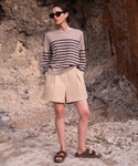 A woman stands outdoors on rocky ground, hands in pockets, wearing sunglasses, a beige and black striped sweater, beige shorts, and the Suede Hutton Slide Sandal—the perfect warm-weather shoe—while looking to the side.