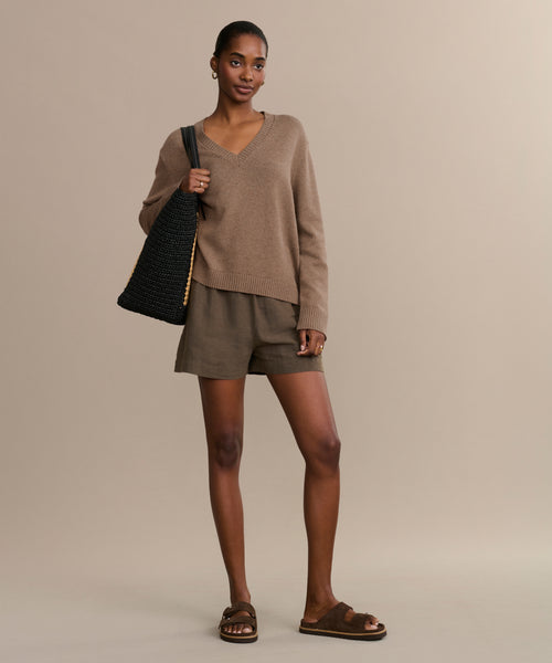 A woman stands against a beige background in a brown V-neck sweater, brown shorts, hoop earrings, a black textured shoulder bag, and completes her look with chic Suede Hutton Slide Sandals.