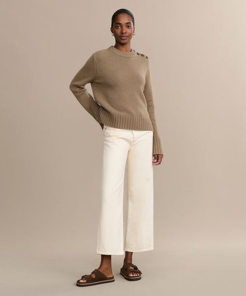 A woman stands against a neutral background in a tan sweater with shoulder buttons, wide-leg off-white pants, and Suede Hutton Slide Sandals. She has her hand on her hip and wears a neutral expression.