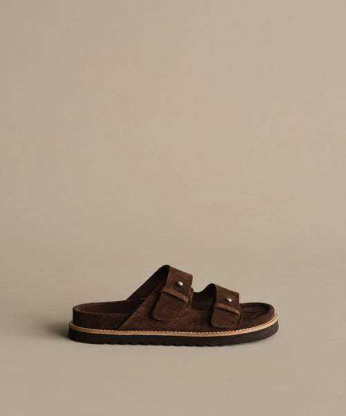 The Suede Hutton Slide Sandal, shown in profile against a beige background, features brown suede, two adjustable buckle straps, and a flat sole—perfect for warm-weather style.
