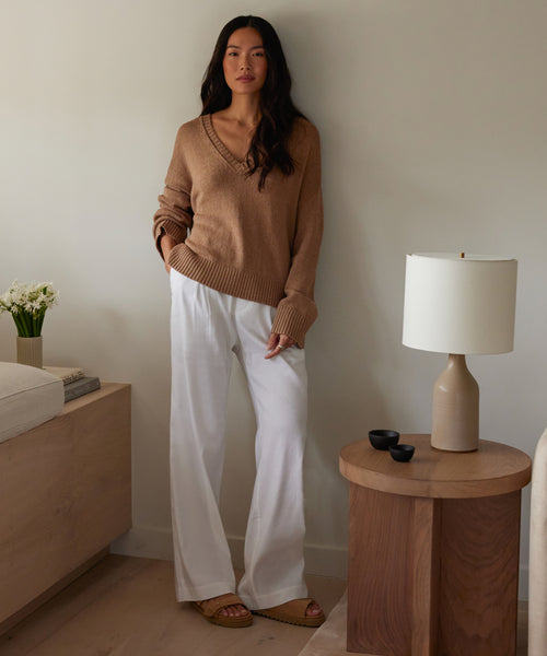 A woman stands by a wooden table, wearing a brown sweater, white wide-leg pants, and the Suede Hutton Slide Sandal—a stylish warm-weather choice. The room has neutral decor: a lamp, small bowls, flower vase, and stacked books.