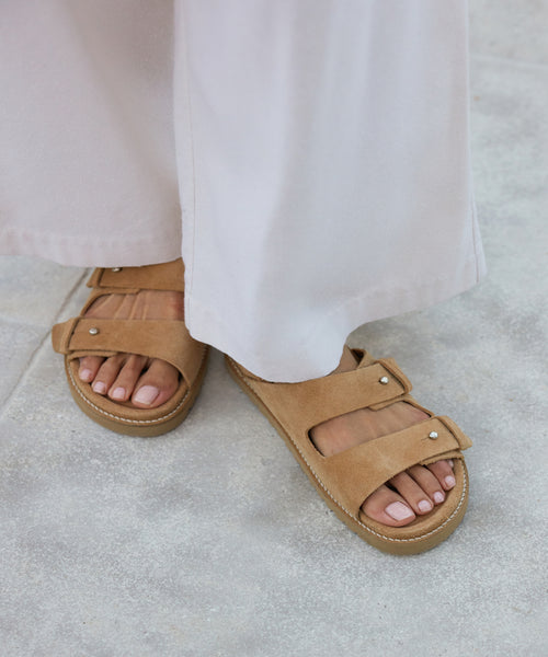 A person wearing wide-legged light pants and the Suede Hutton Slide Sandal with signature push-button details stands on a light gray textured surface, their neatly manicured toes highlighting this ideal warm-weather shoe.