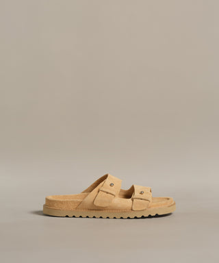 The Suede Hutton Slide Sandal, a tan suede slide with two buckle straps and a ridged sole, is shown against a plain beige background.