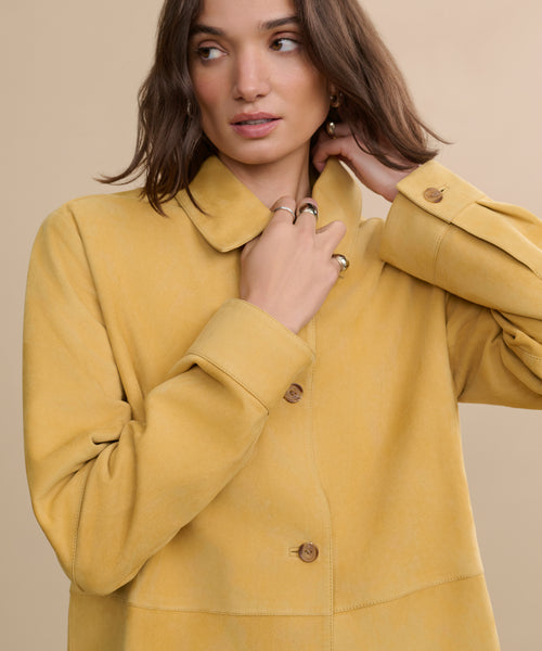 A woman with wavy brown hair wears the Suede Lennox Jacket in a honey shade and gold rings, looking to the side with one hand touching her neck against a beige background.