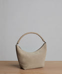 The Medium Crescent Bag in beige suede, made in Italy, features a curved shape and thick twisted handle. It is displayed on a light wooden surface with a plain gray background.
